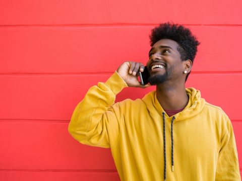Man talking on phone wearing a yellow sweatshirt in front of a pink wall. 