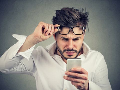 Man holding his glasses over his head and looking confused at his phone. 