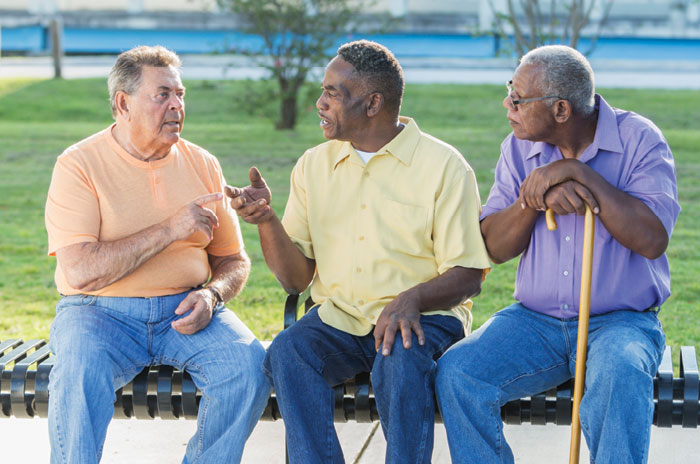 Three men sitting on a bench outside talking. 